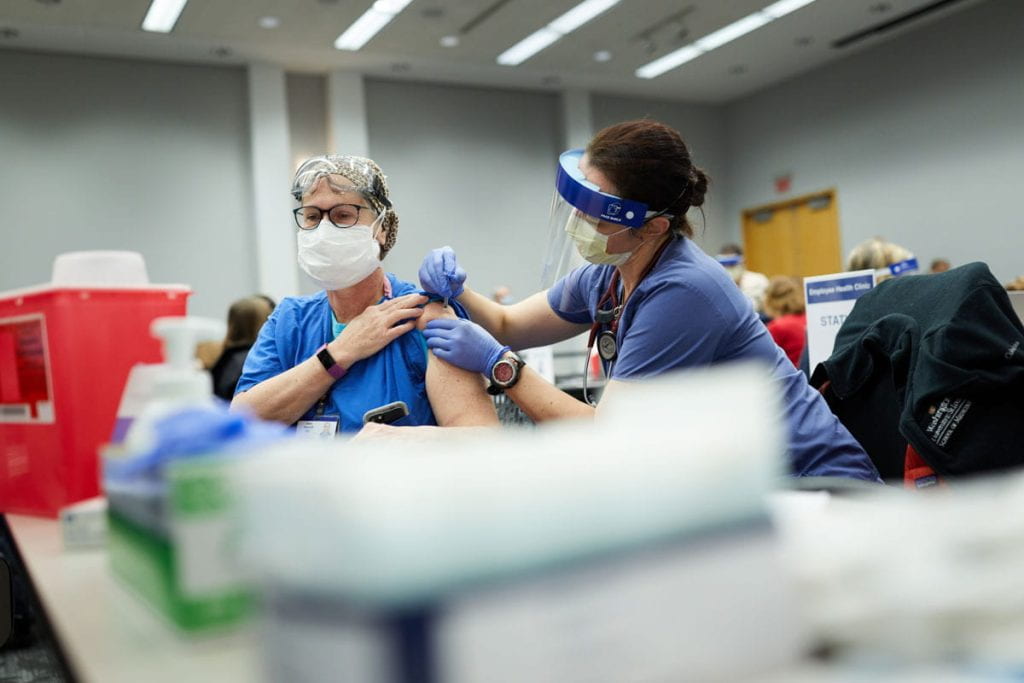 A woman wearing a face shield vaccinates a woman wearing blue scrubs and a mask